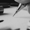 Photo de la main de l'artiste qui tient un critérium au dessus d'une feuille, avec d'autres stylos éparpillés sur la table.