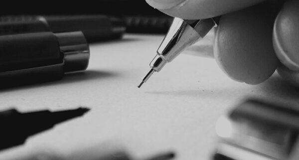 Photo de la main de l'artiste qui tient un critérium au dessus d'une feuille, avec d'autres stylos éparpillés sur la table.