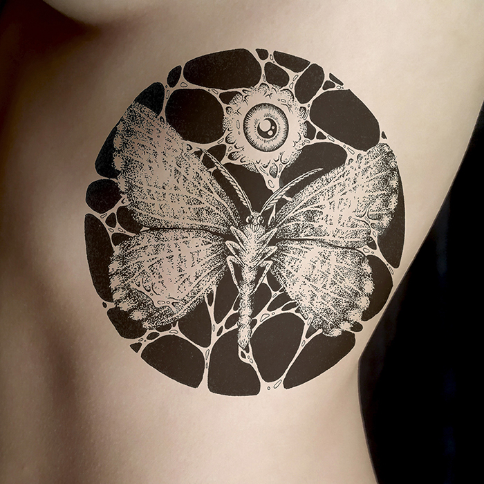 Photo d'une simulation photo d'une illustration tatouée sur un torse féminin, l'œuvre est composée d'un papillon et d'un œil tissés dans un cercle, par l'artiste Okograph.